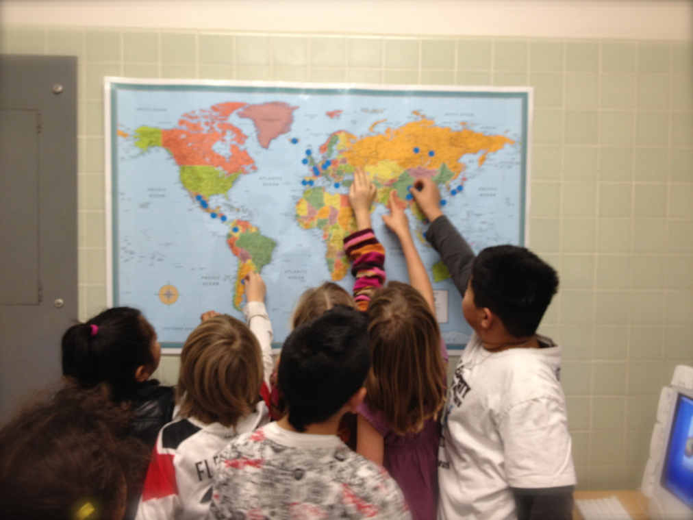 The children point at a map of the world marked with blue sticker dots.